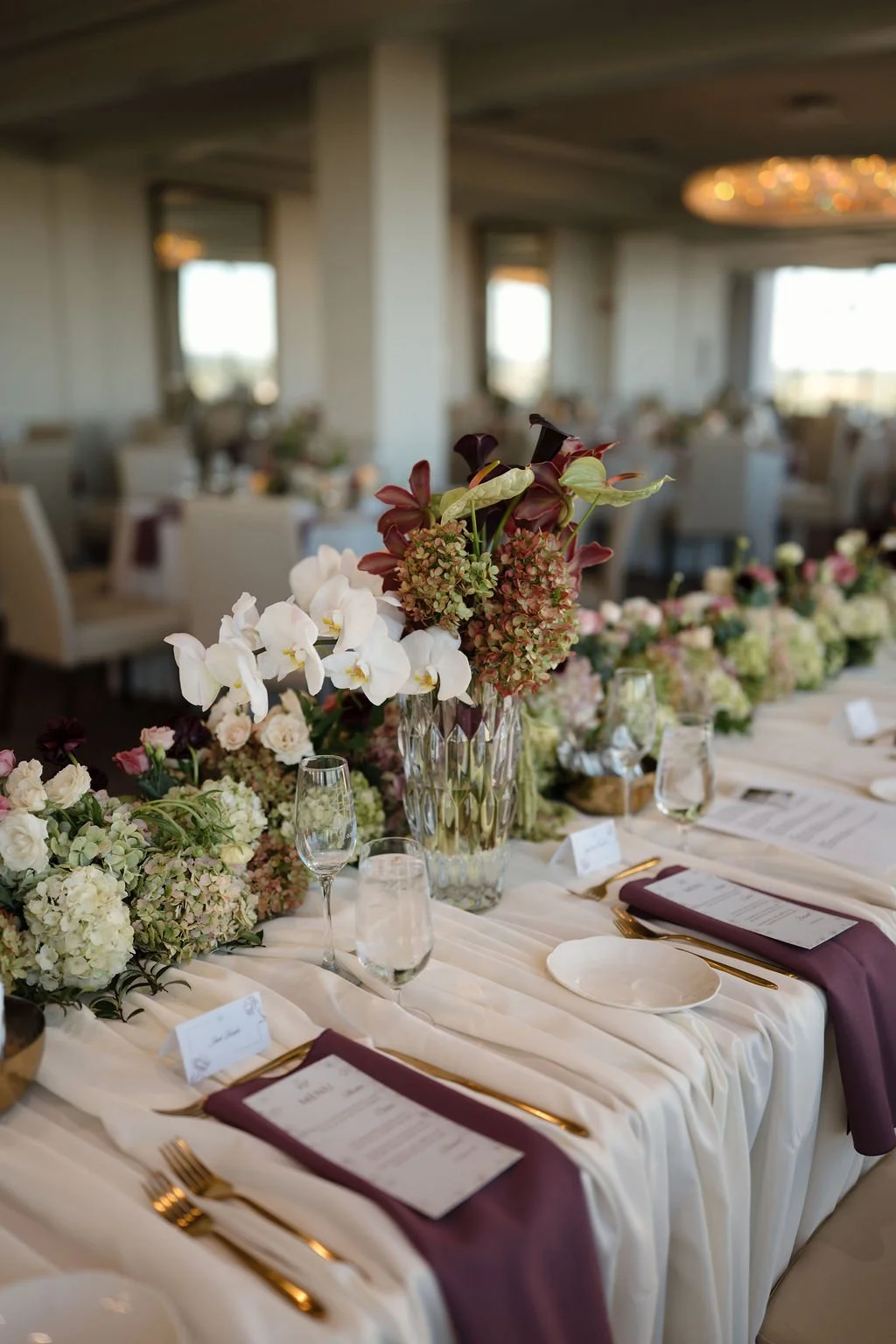 Head table tablescape with white orchids, green hydrangea, and burgundy anthurium in a crystal vase, with plum napkins and printed menus, a romantic detail from Fort Wayne wedding venues