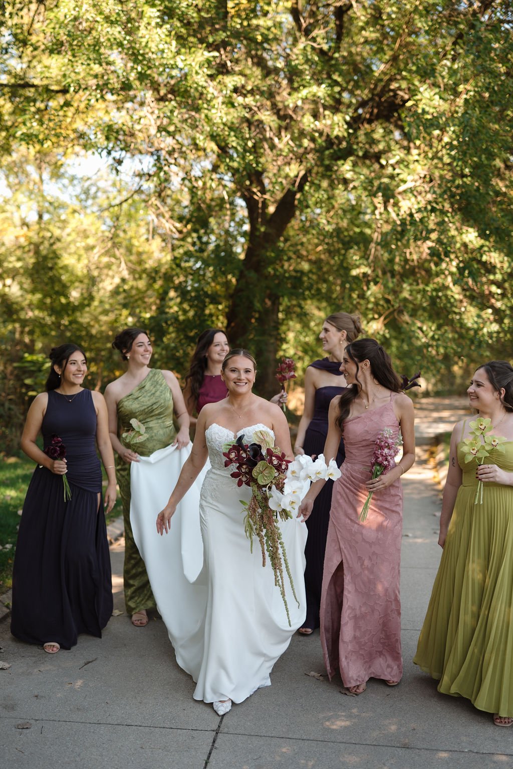 Bride walking with her bridesmaids in mixed jewel-tone gowns in plum, olive, and dusty rose along a tree-lined path