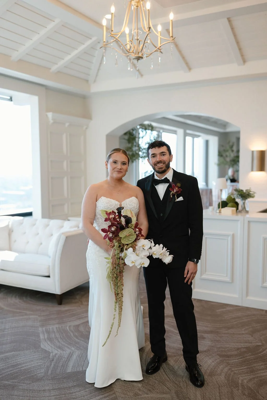 Bride and groom posing together under a gold chandelier, holding a cascading bouquet of burgundy orchids, white phalaenopsis, and green amaranthus