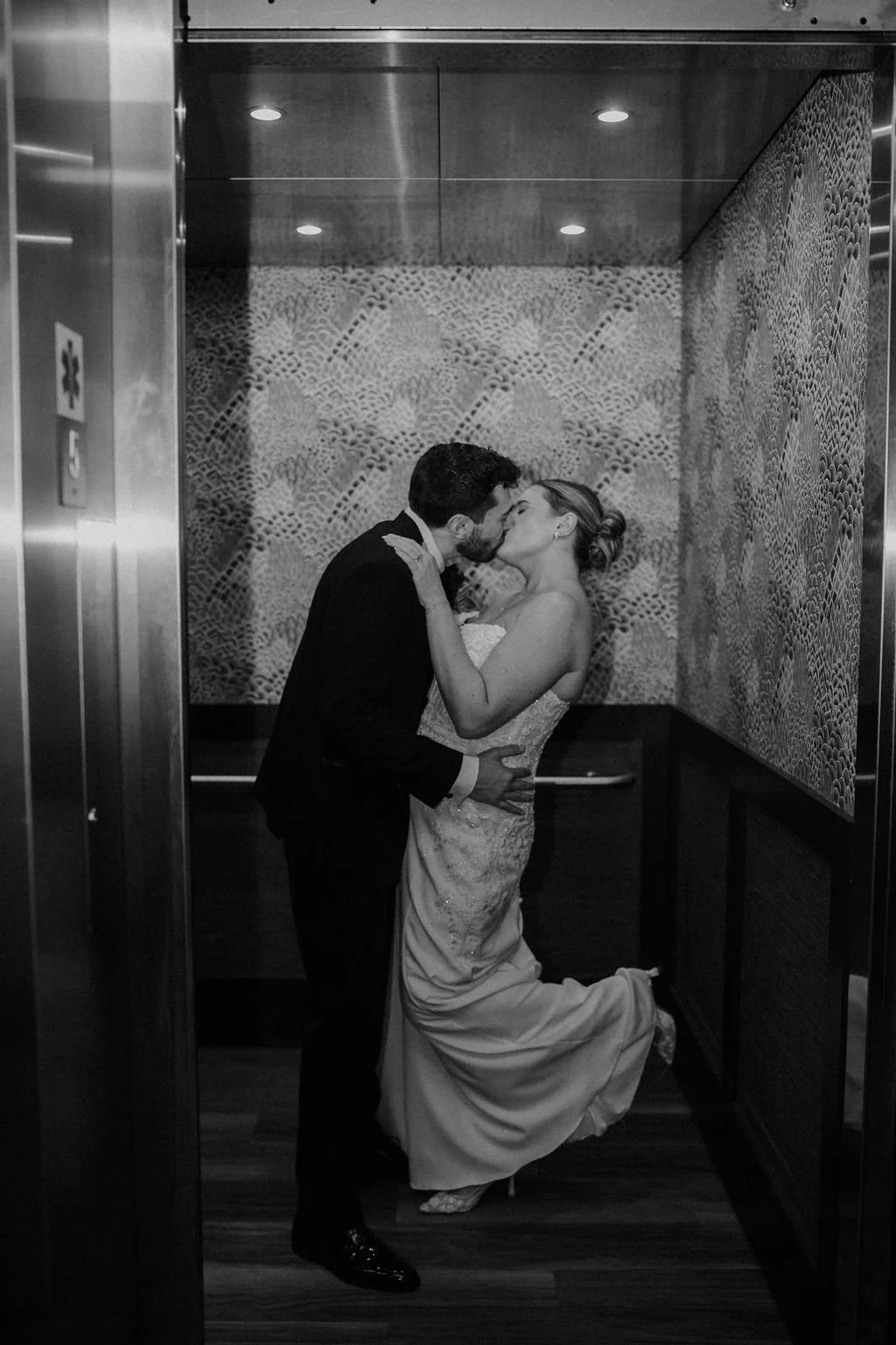 Black and white photo of a bride and groom kissing in an elevator with patterned wallpaper as the bride kicks up her heel