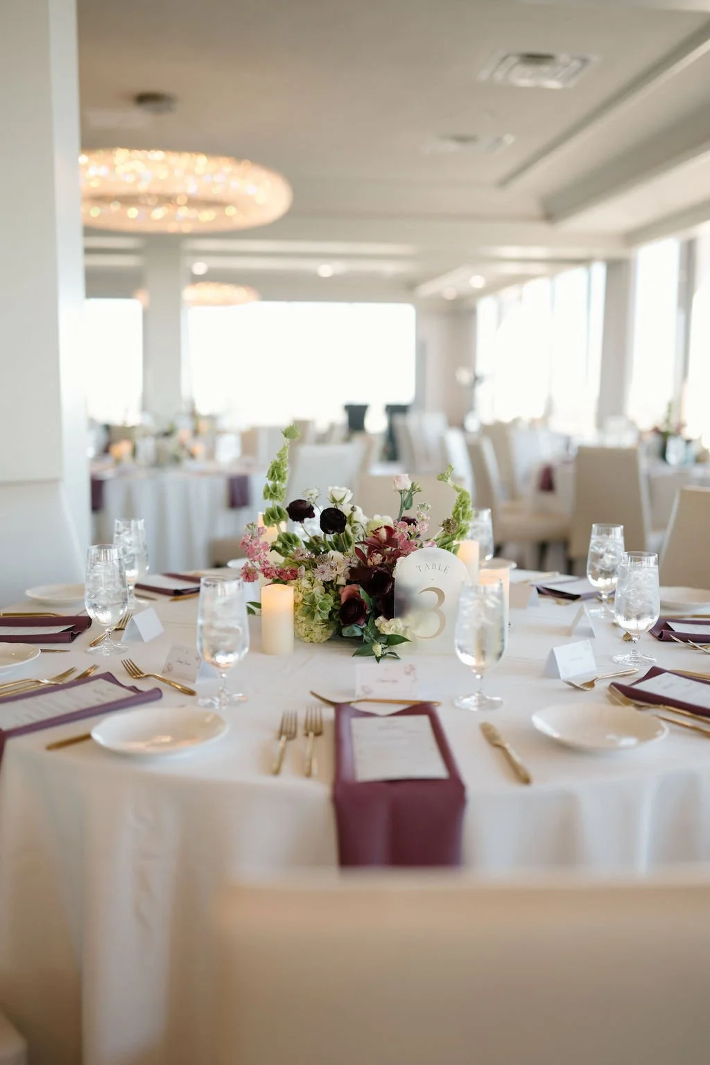 Round reception table with a low floral centerpiece of burgundy ranunculus and green hydrangea, and plum napkins with gold flatware, a polished tablescape from one of the Fort Wayne wedding venues