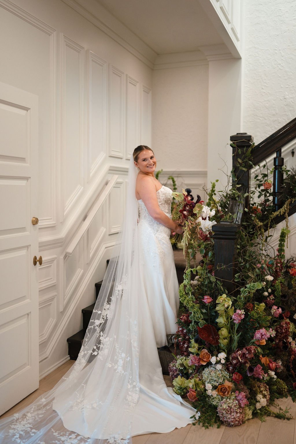 Bride in a strapless lace gown and long floral lace veil standing on a staircase beside a lush wild floral installation
