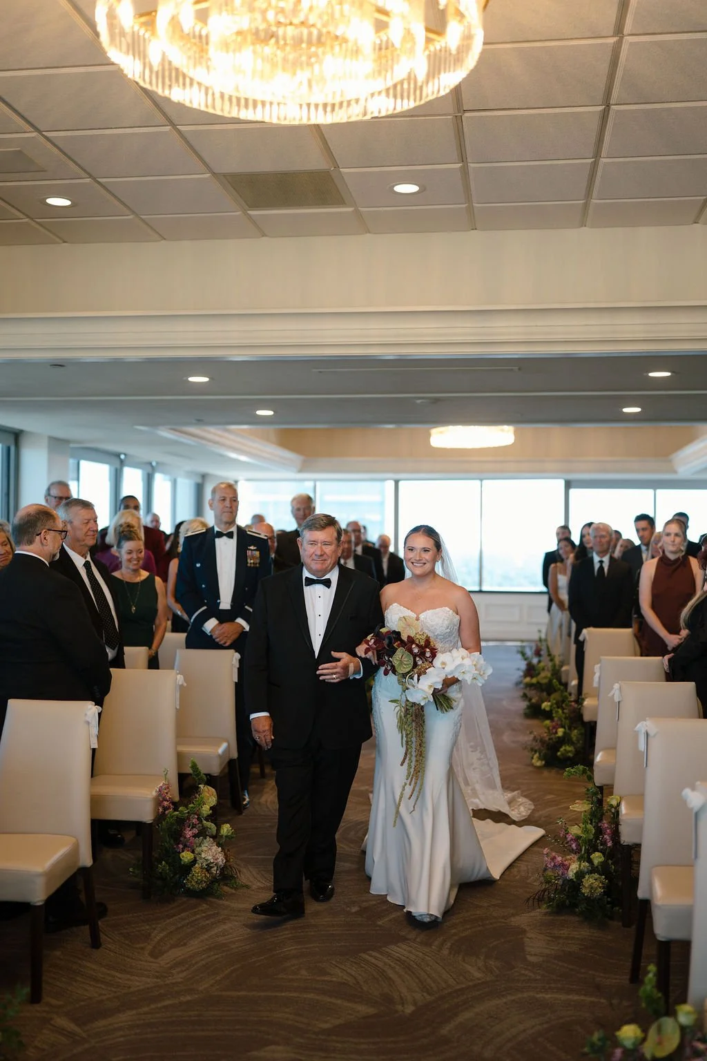 Bride smiling as her father walks her down the aisle past rows of guests under a large crystal chandelier