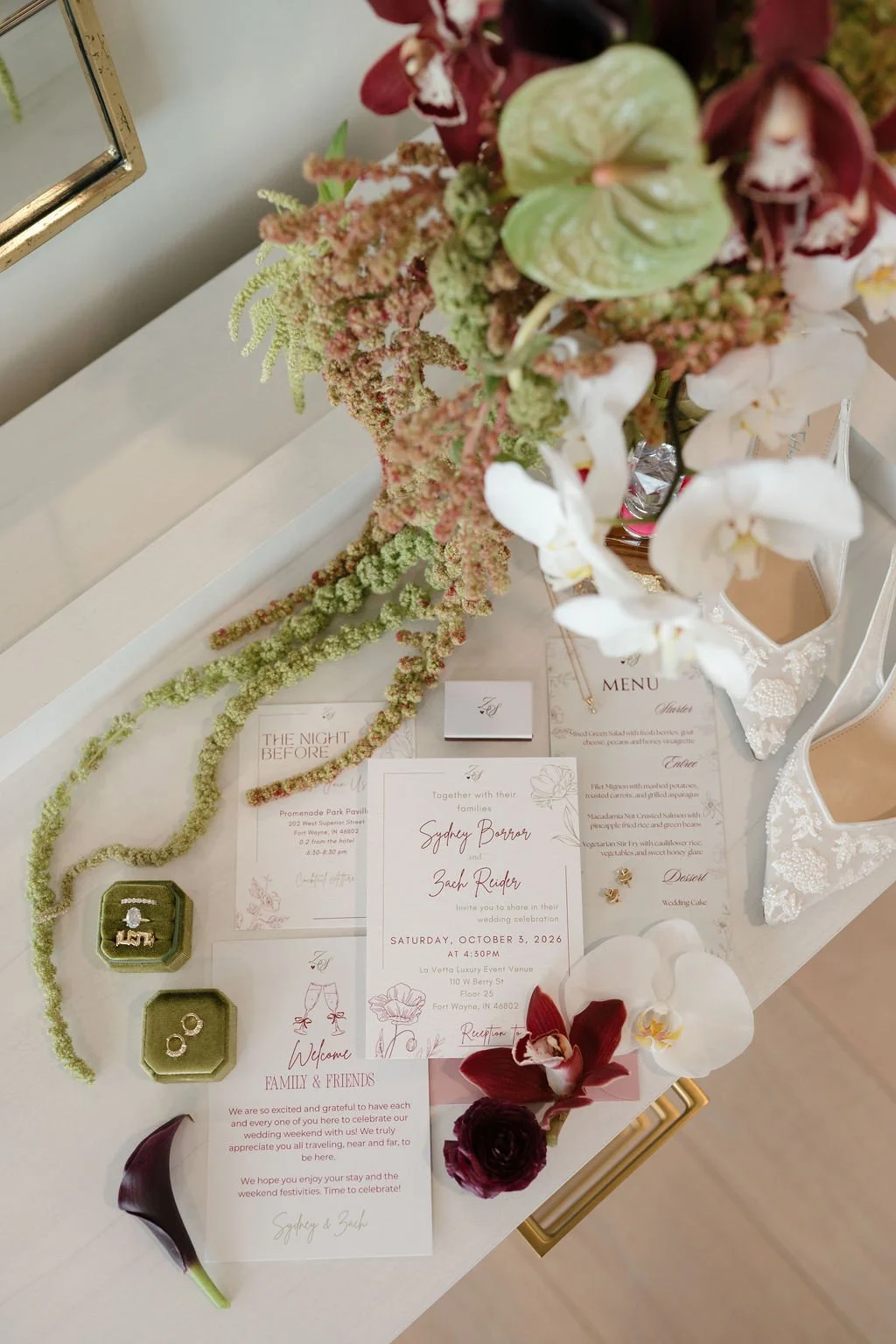 Wedding day flat lay with an invitation suite, menu card, welcome note, gold rings in olive velvet boxes, beaded bridal heels, and a bouquet of white orchids and green amaranthus