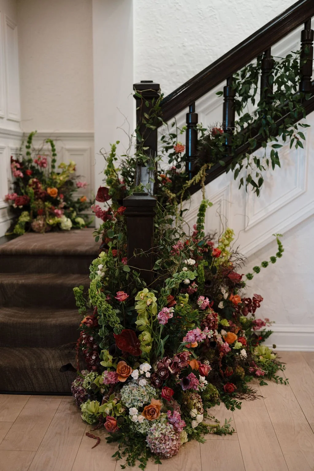 Overgrown floral installation of burgundy, orange, pink, and green blooms wrapped around a dark wood staircase railing, a dramatic design idea for Fort Wayne wedding venues with grand staircases