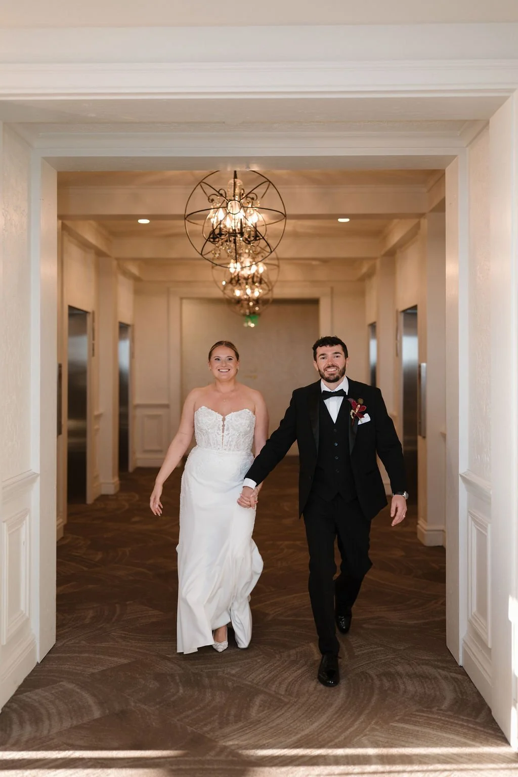 Bride and groom walking hand in hand and smiling down a hallway lined with elevators and orb chandeliers