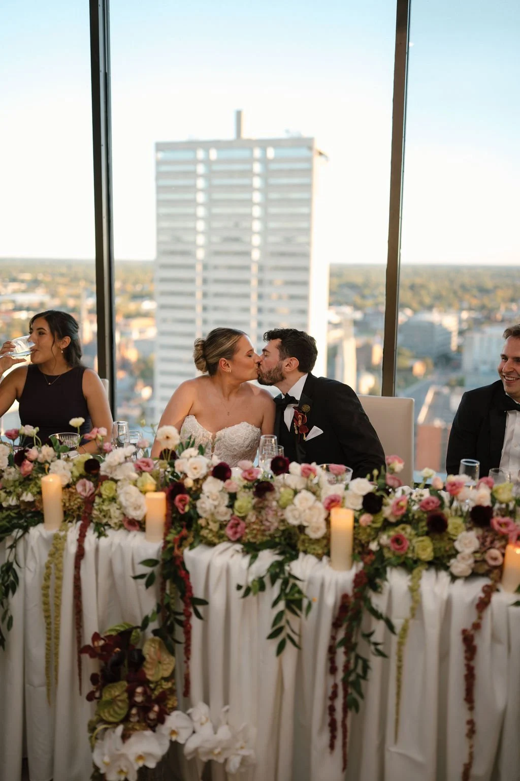 Bride and groom sharing a kiss at the head table, framed by a lush floral garland, pillar candles, and city skyline views, proving why rooftop Fort Wayne wedding venues are so romantic