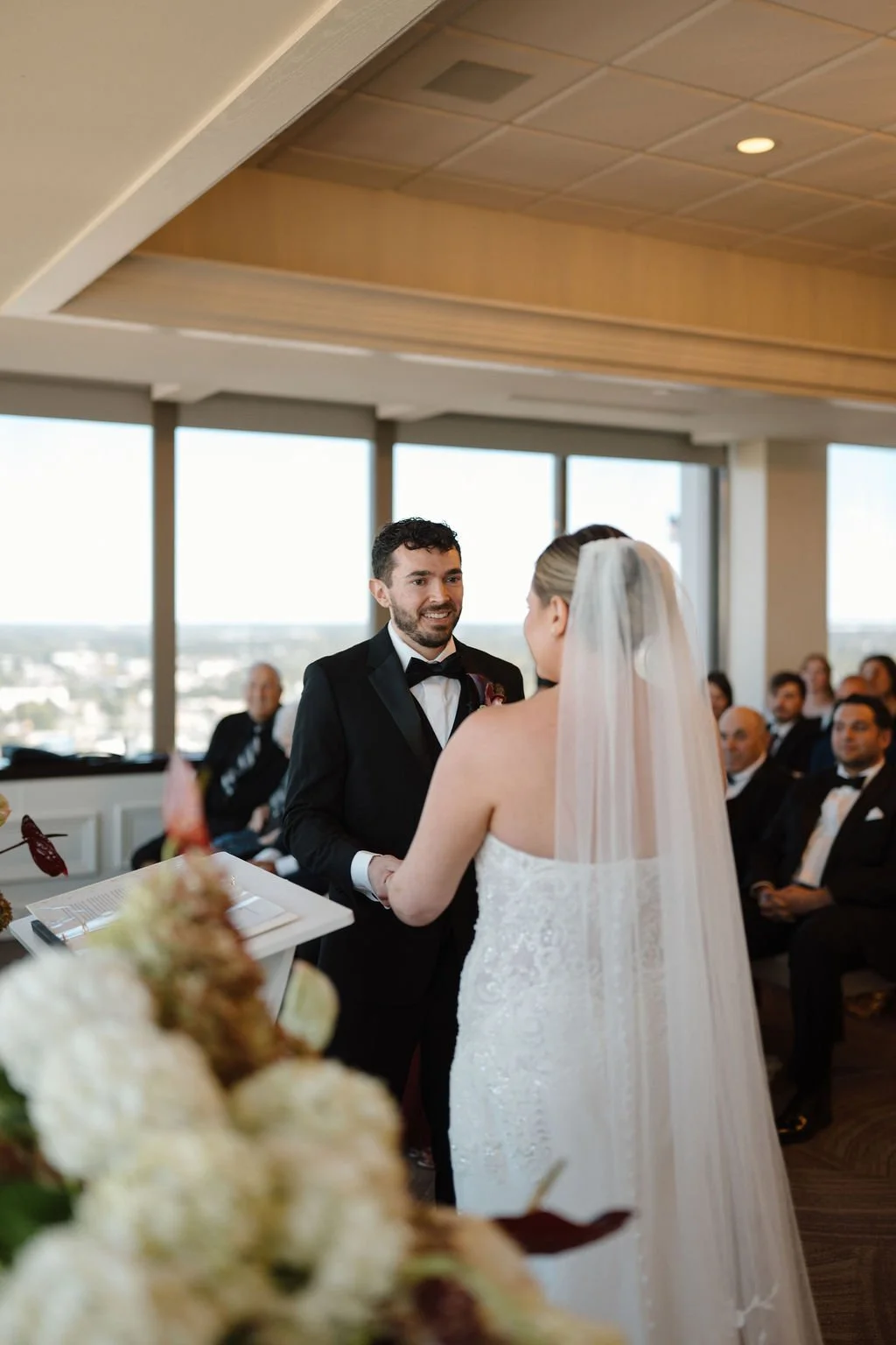 Groom smiling while holding the bride’s hands during the ceremony, with floor-to-ceiling windows and guests in the background at a high-rise Fort Wayne wedding venue