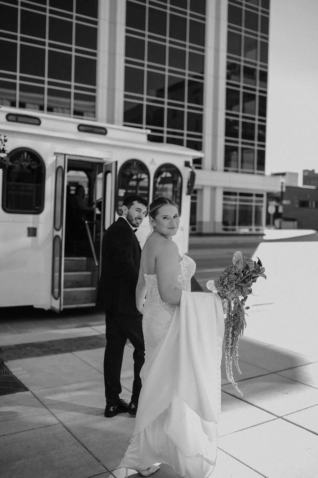 Black and white photo of a bride in a strapless lace gown holding her train and cascading bouquet while looking back with her groom beside a white wedding trolley