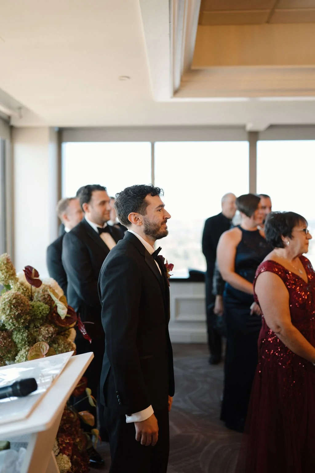 Groom in a black tuxedo and bow tie watching the aisle with an emotional smile as guests stand behind him at the ceremony