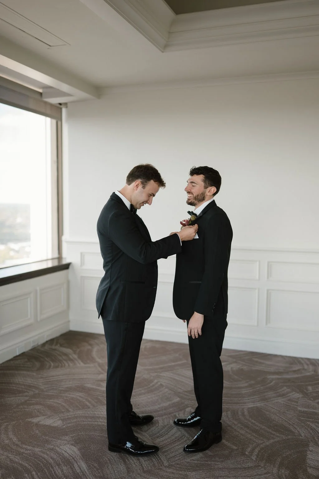 Groomsman pinning a burgundy orchid boutonniere onto the groom’s black tuxedo as both laugh in a bright white paneled room