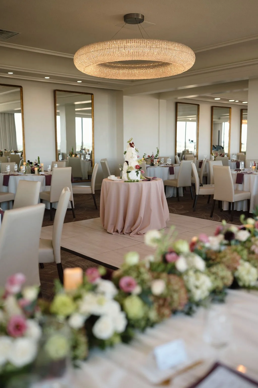 Wedding cake on a blush pink draped table on the dance floor, surrounded by tall gold mirrors, and floral centerpieces, a stunning reception setup at one of the best Fort Wayne wedding venues