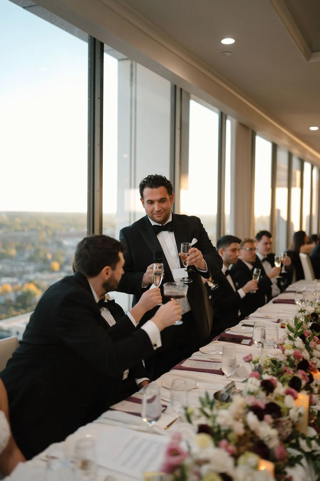 Groomsman standing to toast the head table with a champagne glass and microphone, floral garland in the foreground