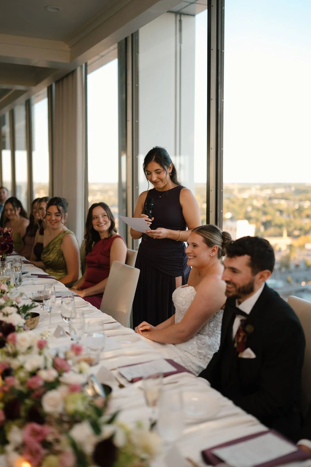 Bridesmaid giving a toast at the head table as the bride and groom smile, with skyline views at one of the Fort Wayne wedding venues