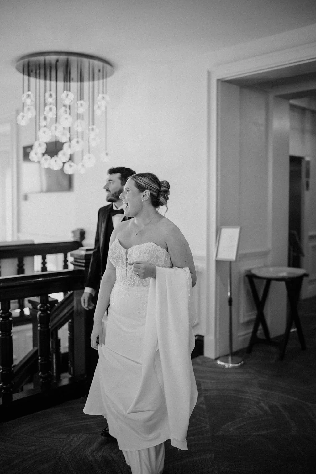Black and white photo of a bride laughing as she walks past her groom, with a modern glass globe chandelier and dark wood staircase railing behind them