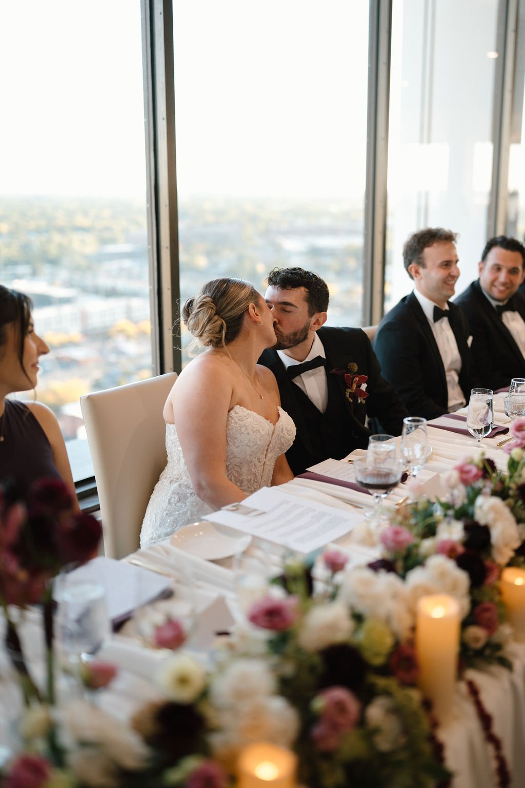 Bride and groom kiss at the head table in front of floor-to-ceiling windows overlooking the city at one of the Fort Wayne wedding venues