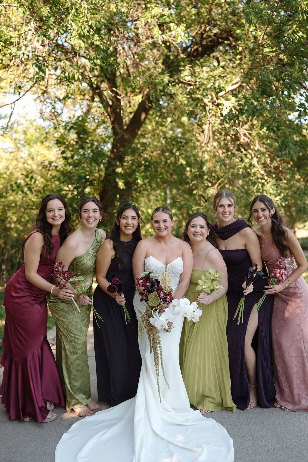 Bride laughing with six bridesmaids in burgundy, olive, navy, and dusty rose gowns under a canopy of green trees