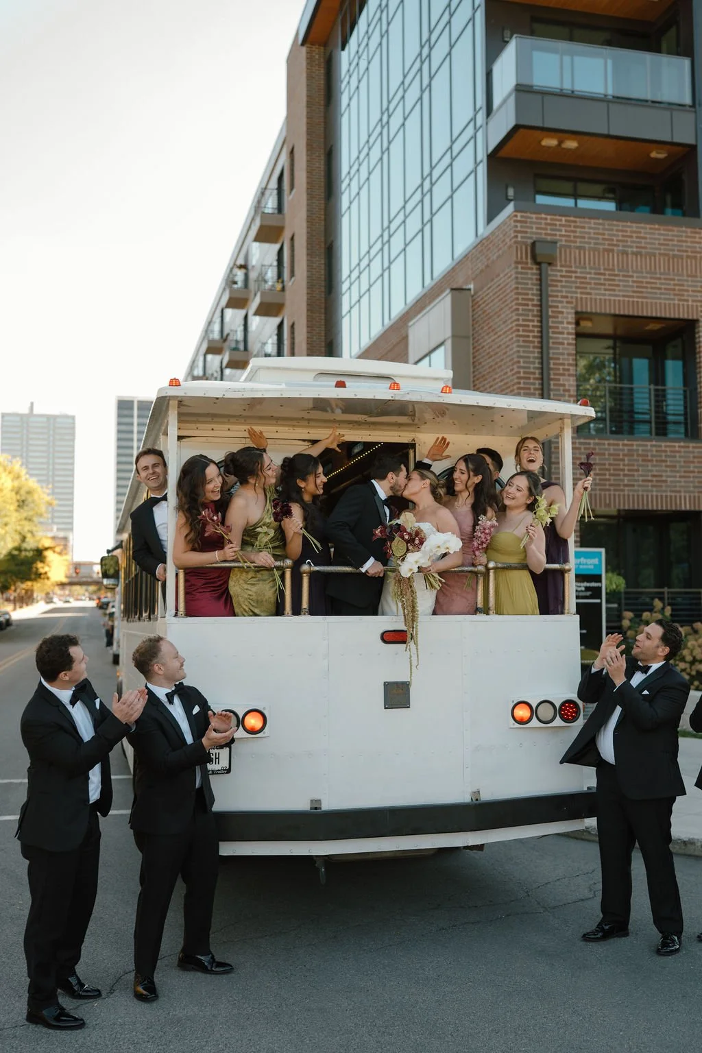 Bride and groom kiss on the back of a white wedding trolley as bridesmaids cheer and groomsmen applaud from the street