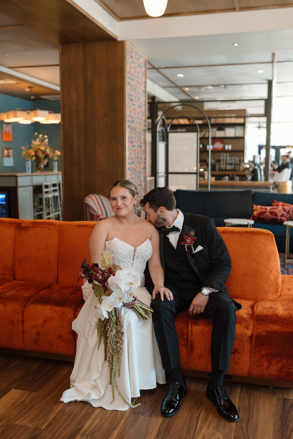 Bride and groom seated on a rust orange velvet sofa in a modern lounge, the groom resting his head on her shoulder