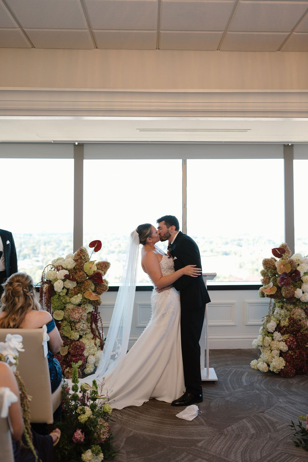 Newlyweds kiss at the ceremony between hydrangea and anthurium arrangements in front of skyline windows at one of the Fort Wayne wedding venues