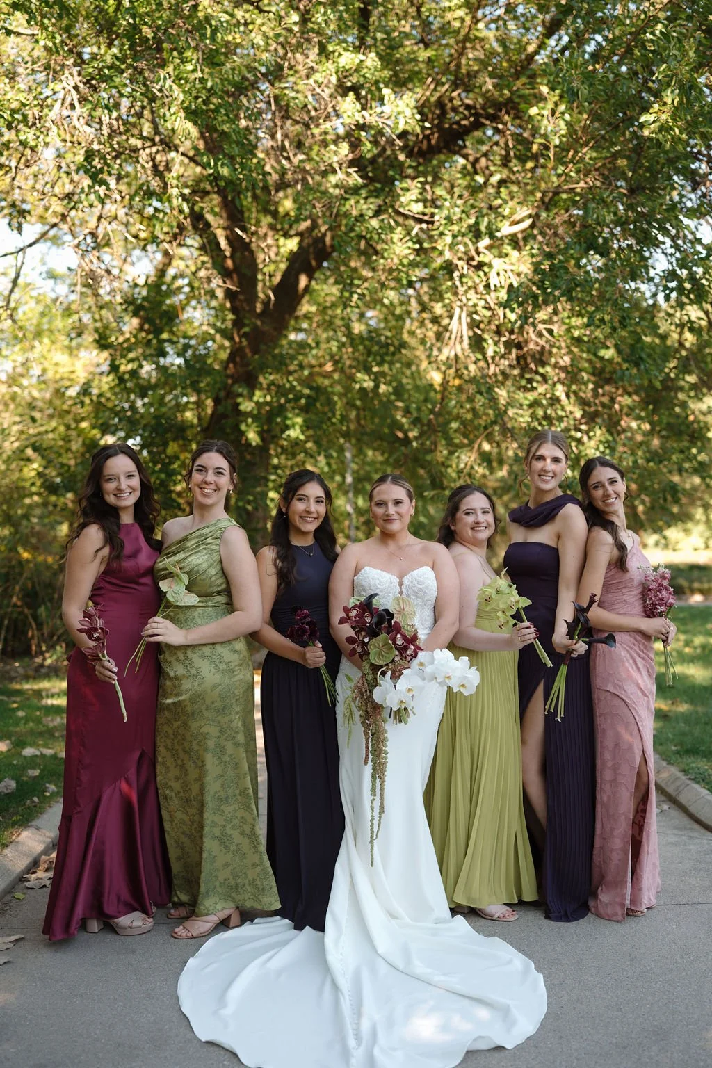 Bride standing with six bridesmaids in mixed burgundy, olive, navy, plum, and dusty rose gowns, each holding a single orchid stem