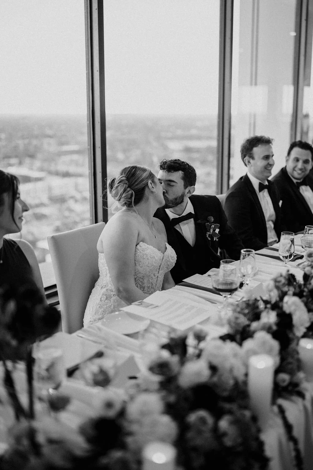 Black and white photo of the bride and groom kissing at the head table with city views and the bridal party smiling nearby