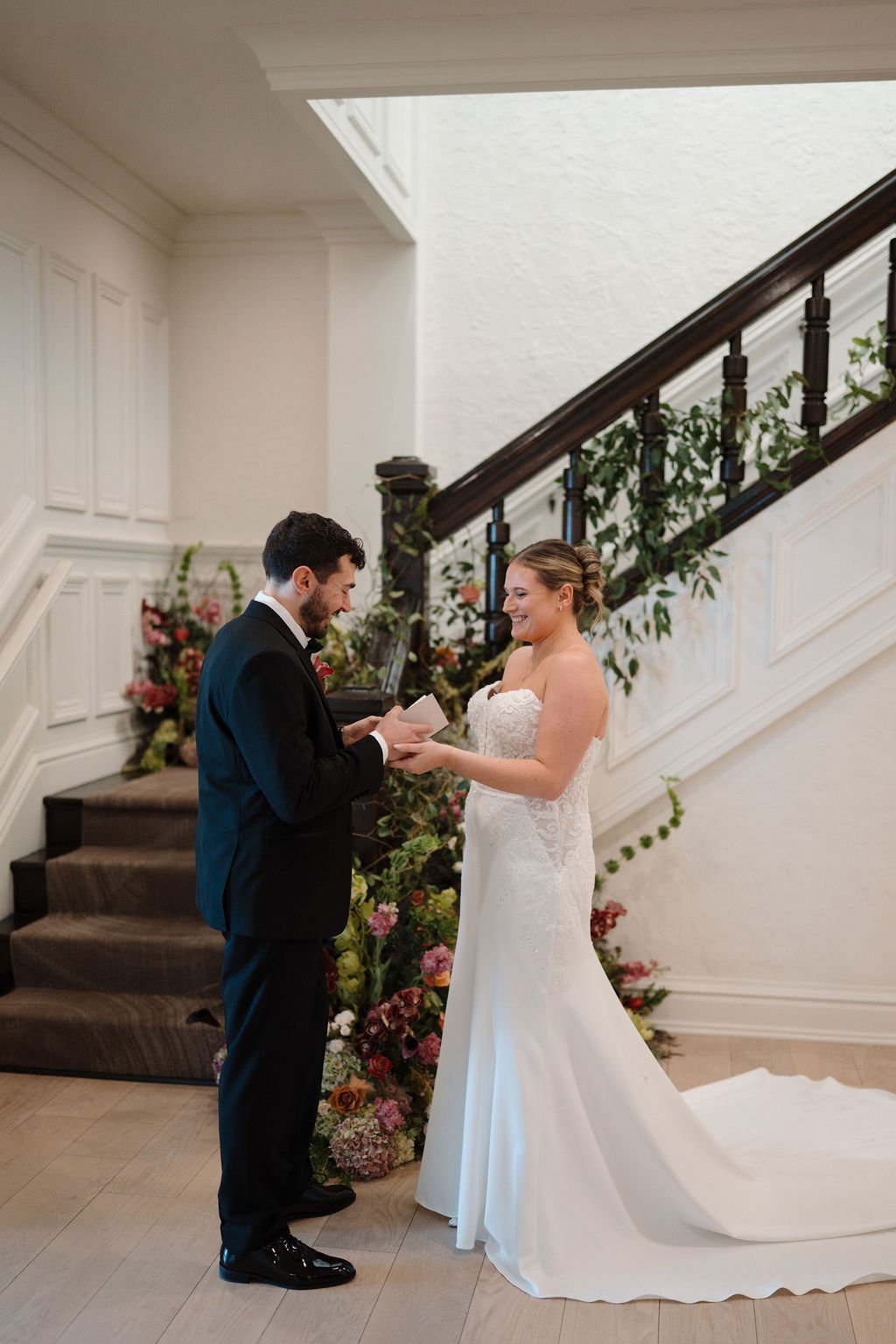Groom reading a note to his laughing bride beside a flower-filled staircase at one of the Fort Wayne wedding venues