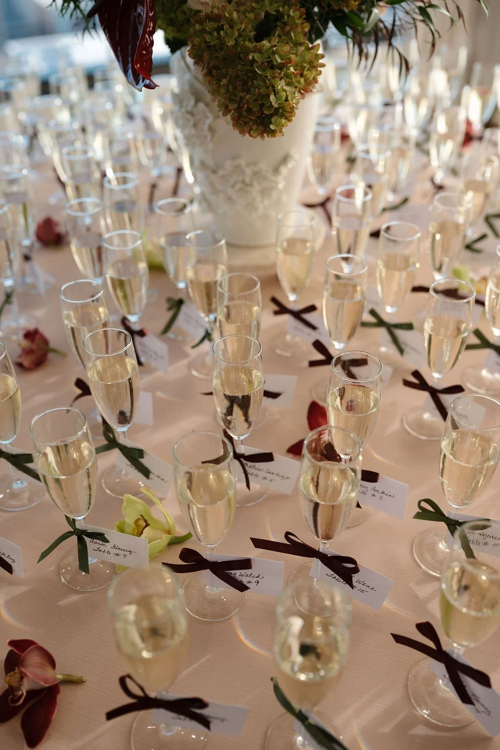 Champagne flutes lined up on a blush table, each tied with a burgundy or green ribbon and a handwritten escort card
