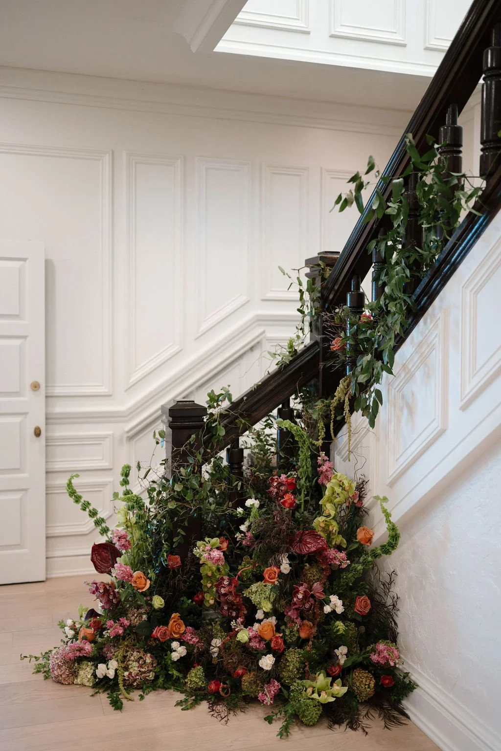 Lush floral installation of burgundy, orange, pink, and green blooms climbing a dark wood staircase railing against white paneled walls