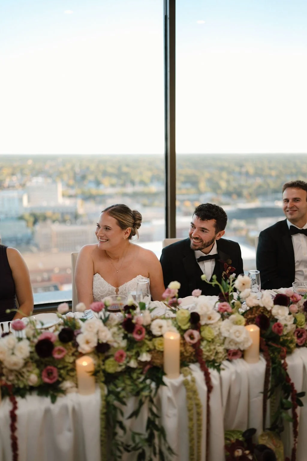 Bride and groom smiling at the head table behind a lush floral garland and pillar candles, with a skyline view at one of the Fort Wayne wedding venues