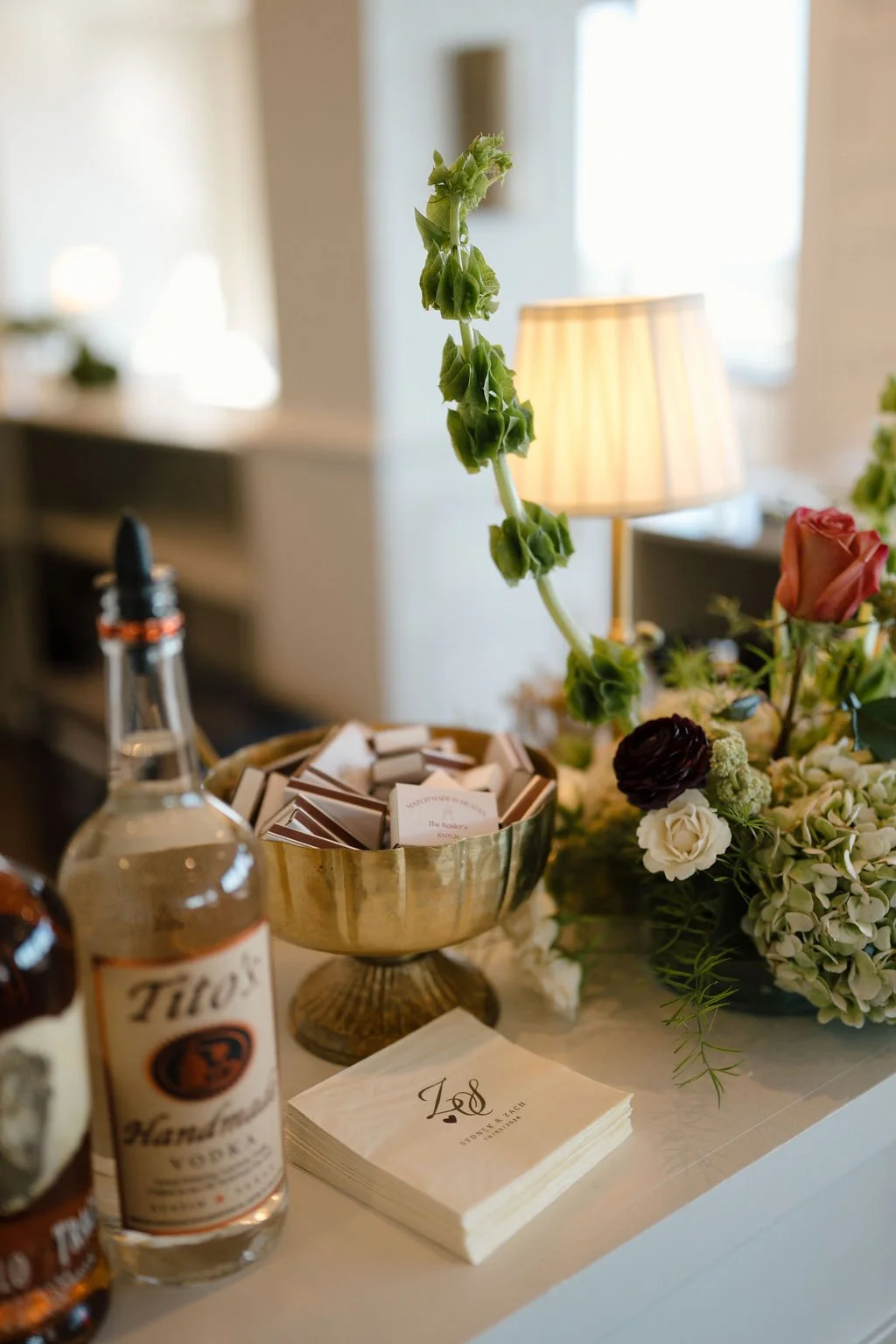 Personalized wedding napkins and a gold bowl of custom matchbooks beside a bottle of vodka and a bell of Ireland floral arrangement at the bar