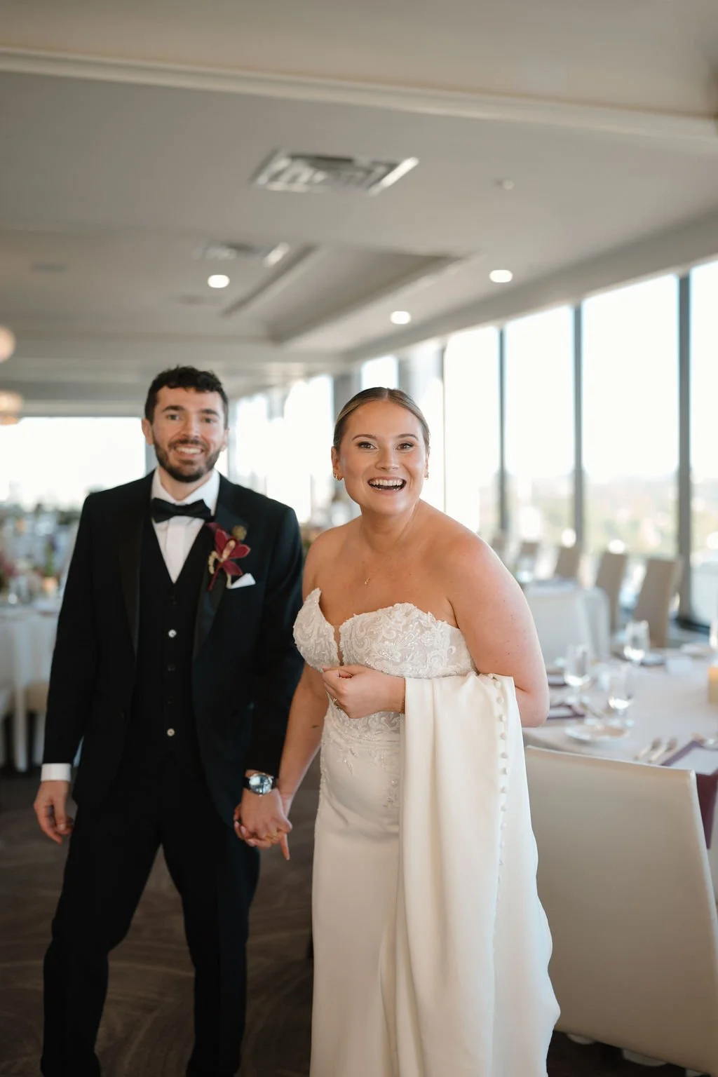 Bride and groom walking hand in hand through the reception room and laughing, with floor-to-ceiling windows and set tables behind them