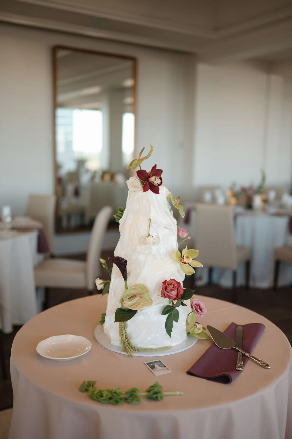 Sculptural white wedding cake decorated with burgundy orchids, pink roses, and anthurium on a blush table at one of the Fort Wayne wedding venues