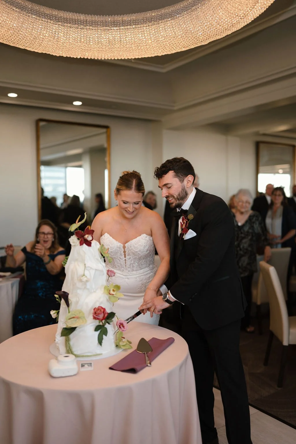 Bride and groom smiling as they cut their sculpted white wedding cake with fresh orchids and roses while guests cheer behind them under a crystal chandelier