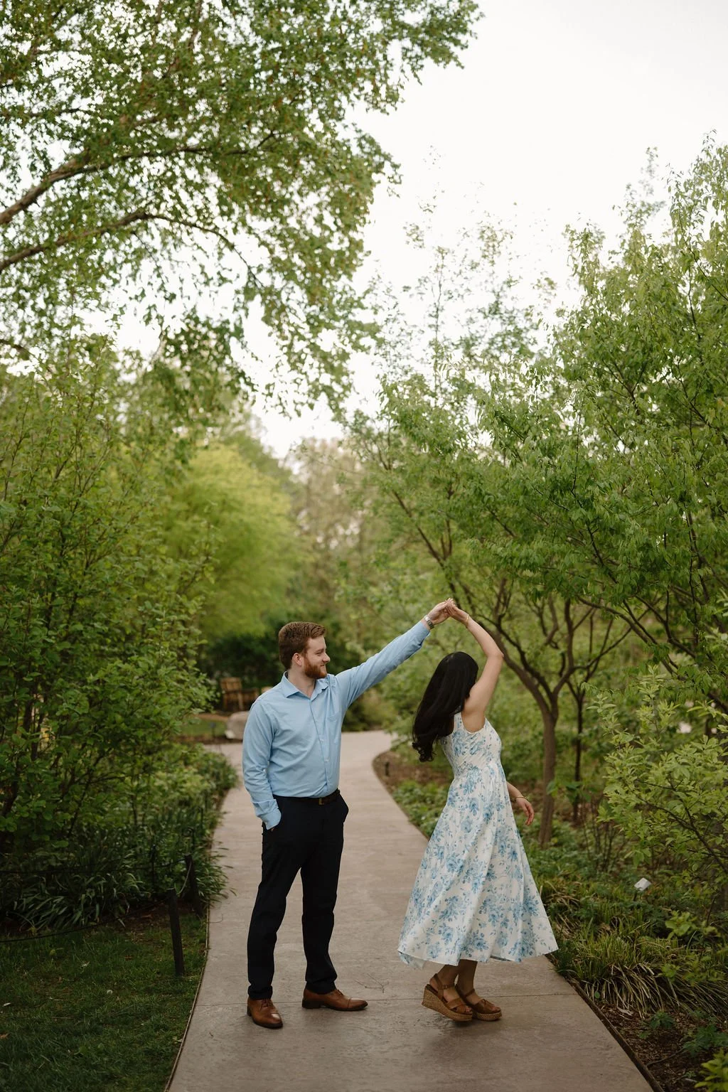 Couple dancing on a tree lined garden path as he twirls her in a blue floral dress.