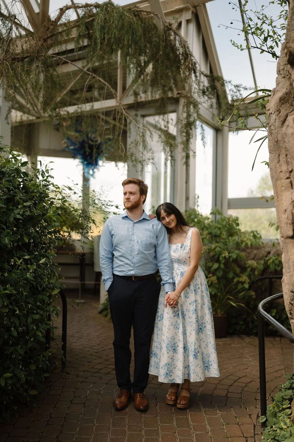 Woman in a blue floral dress resting her head on her fiancé’s shoulder as they stand together on a brick path inside a greenhouse.