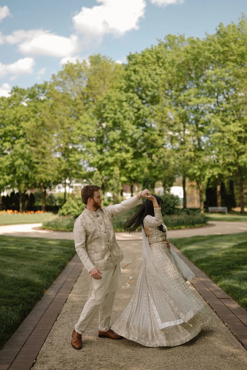 Couple in ivory outfits dancing on a garden path, with his arm raised as she spins and her sequin skirt flares out.