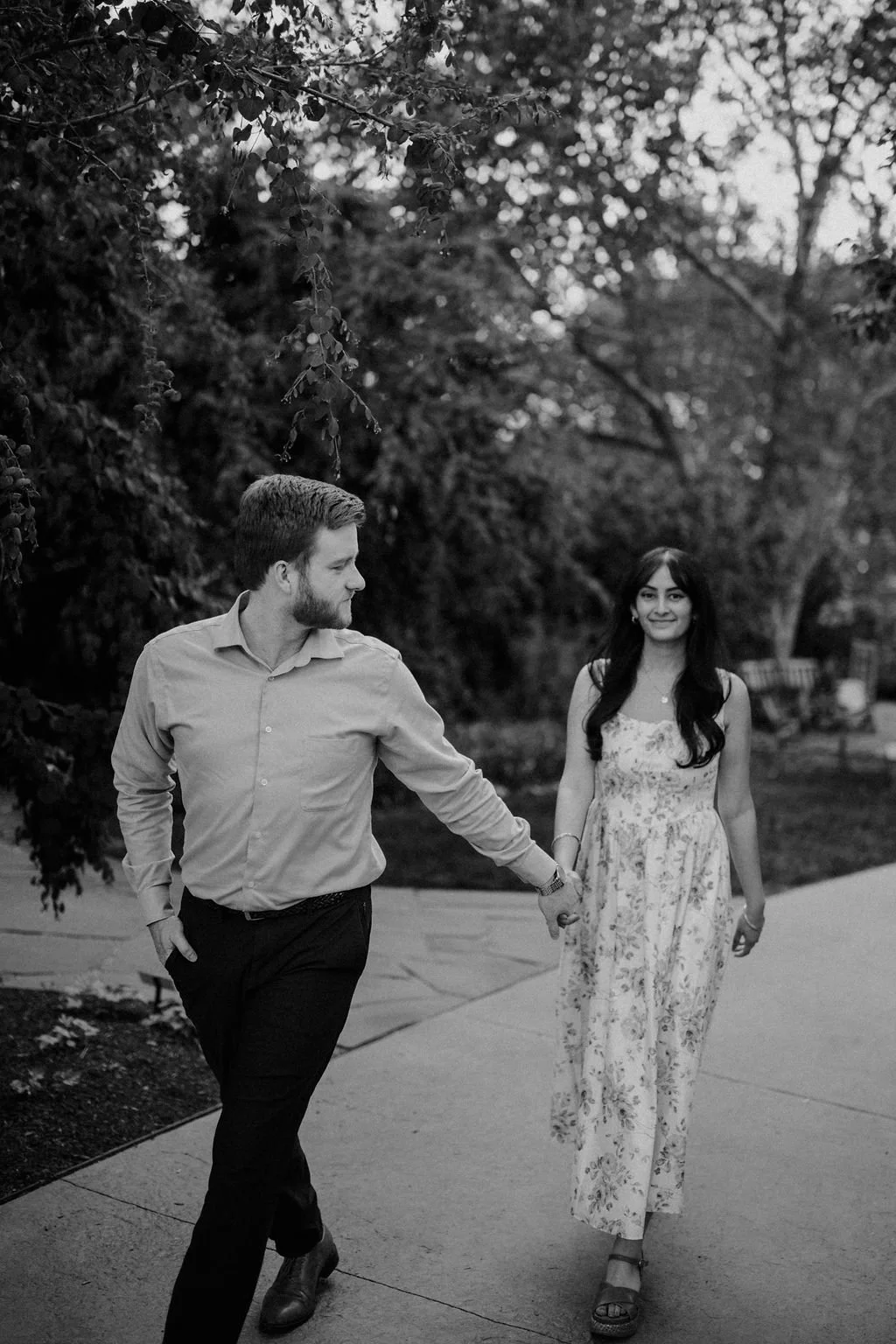 Black and white photo of a man glancing back as he leads his smiling fiancée by the hand along a garden path.