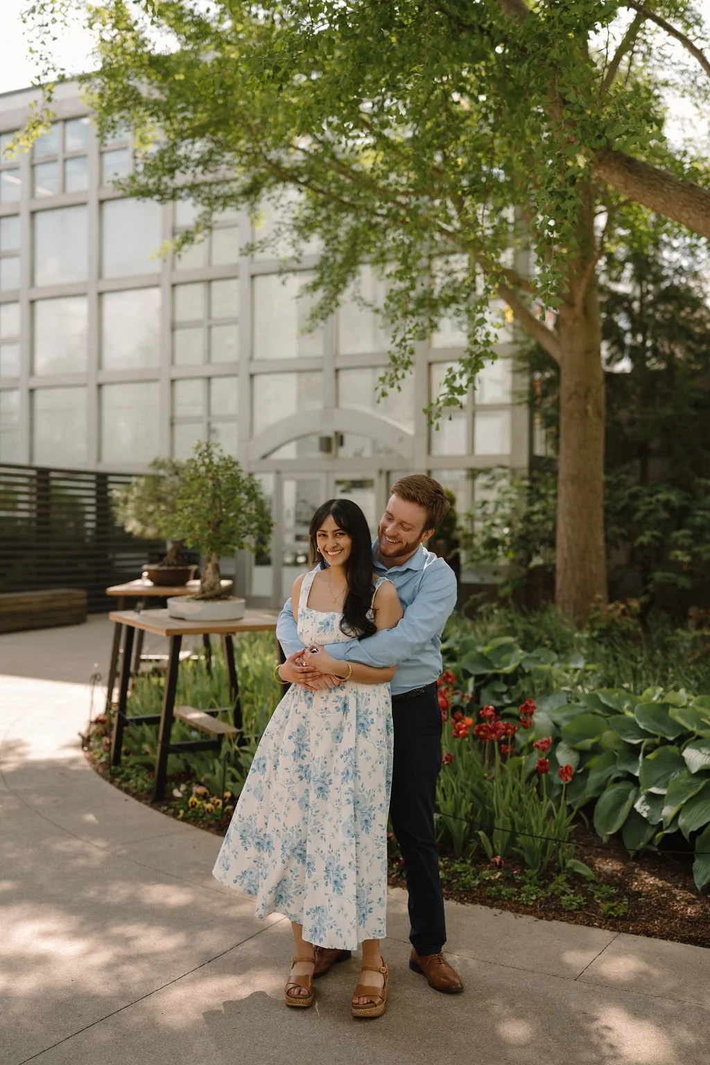 Smiling woman in a blue floral dress being hugged from behind by her fiancé under a ginkgo tree, with red tulips in the garden beside them.