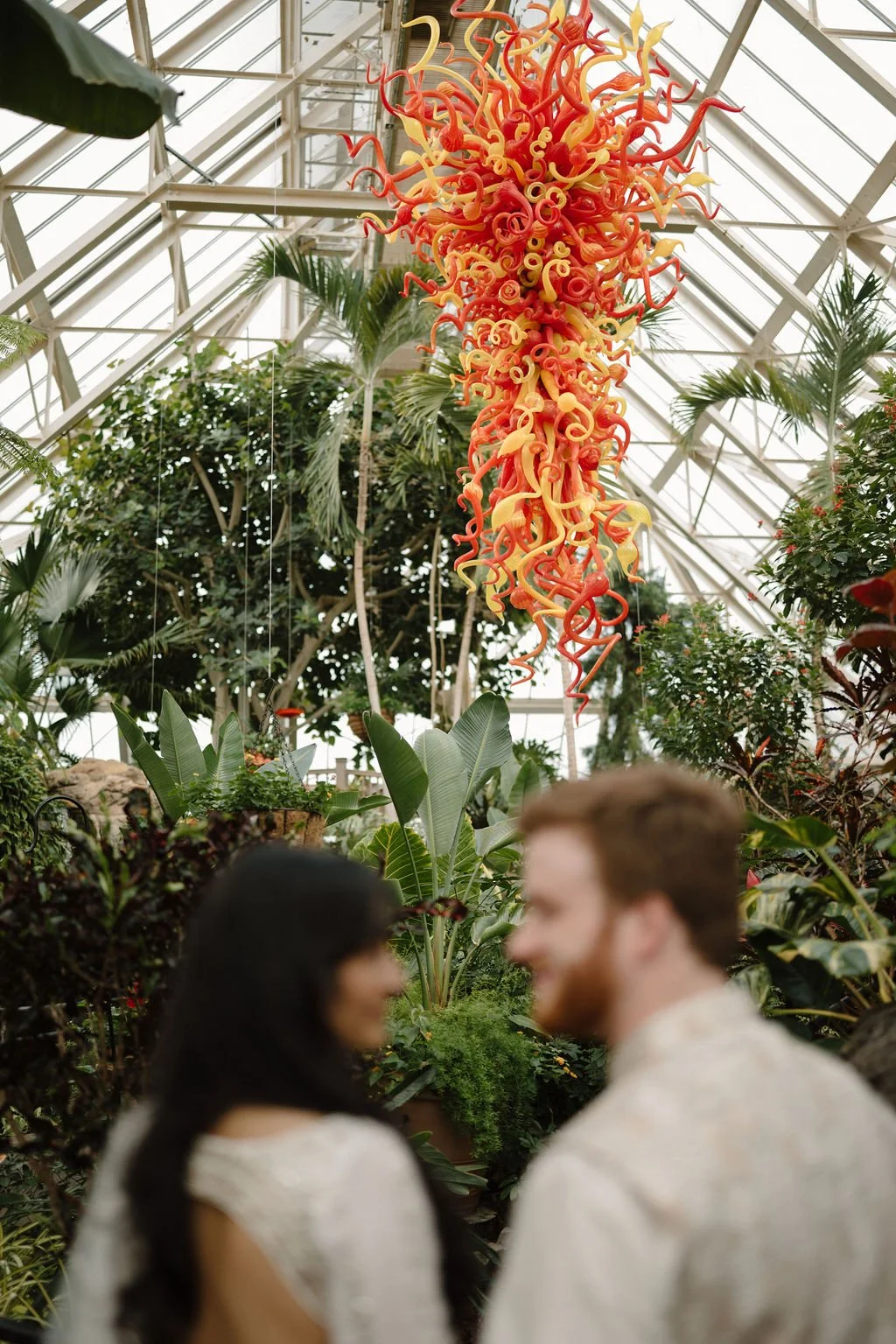 Orange and yellow blown glass sculpture hanging above the plants in a greenhouse, with a couple softly blurred in the foreground.