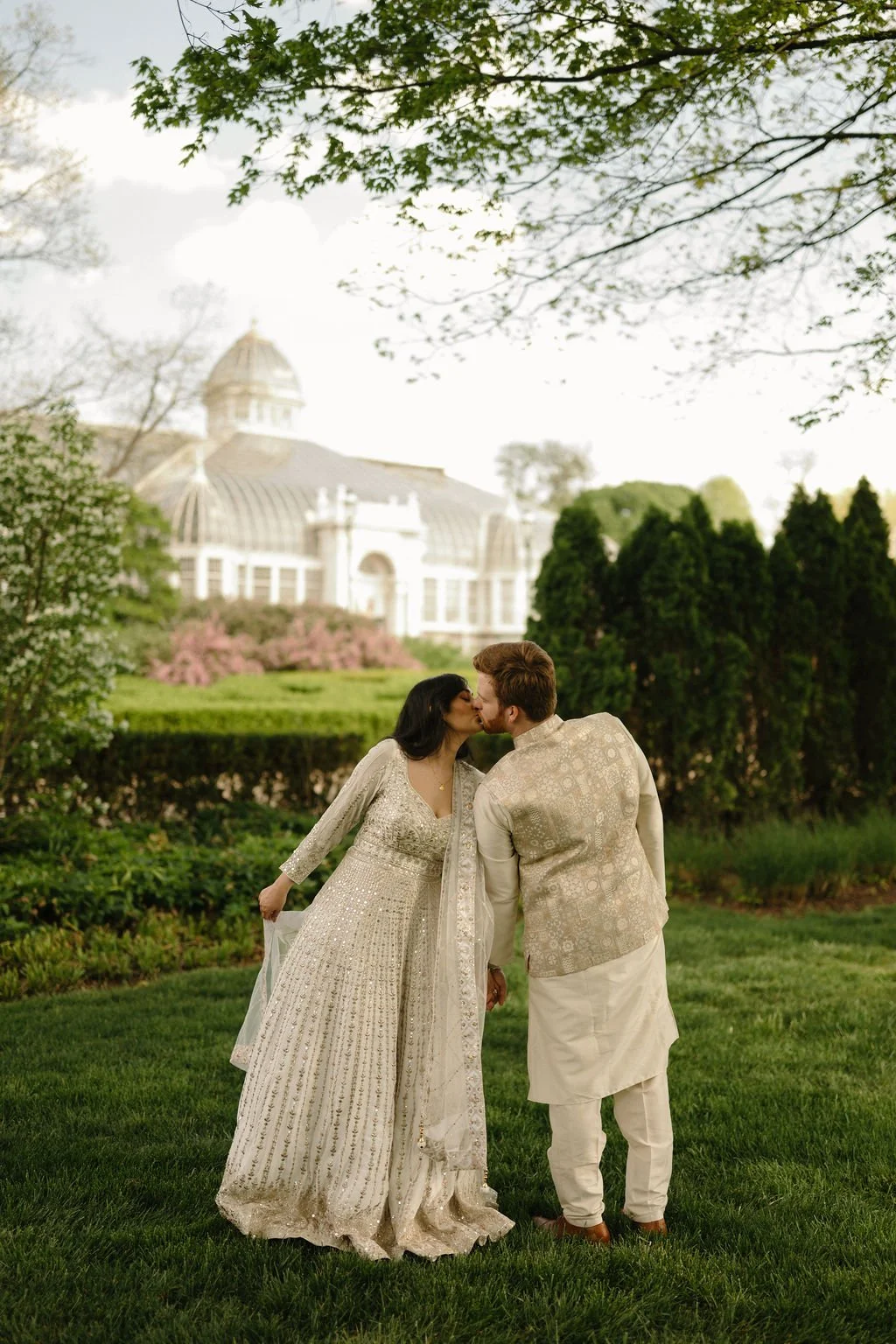 Couple in ivory and gold outfits sharing a kiss on the grass while she holds out her dupatta, with a glass domed conservatory in the background.