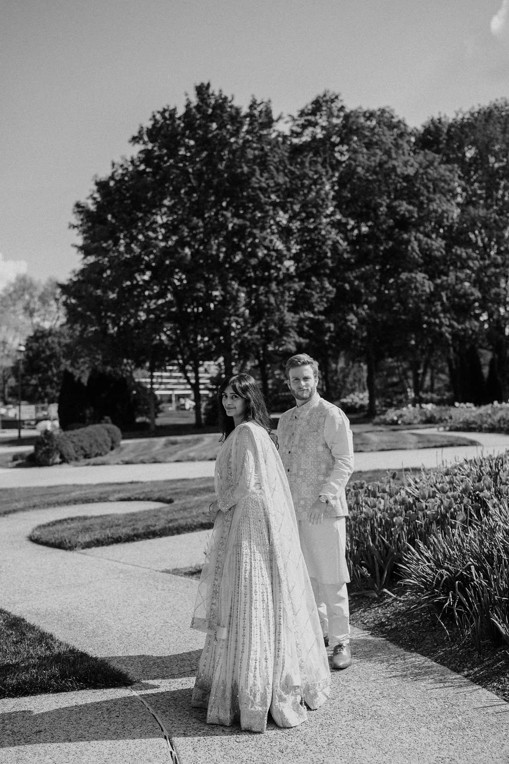 Engagement photo ideas in black and white, with a woman in an ivory sequin skirt looking back over her shoulder as her fiancé stands beside her on a garden path.