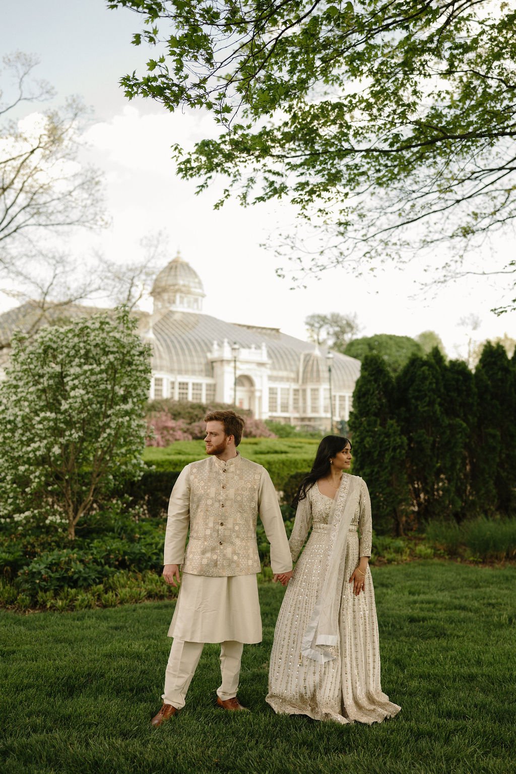 Engagement photo ideas featuring a couple in coordinating ivory outfits holding hands on a lawn, with a glass conservatory and blooming trees behind them.