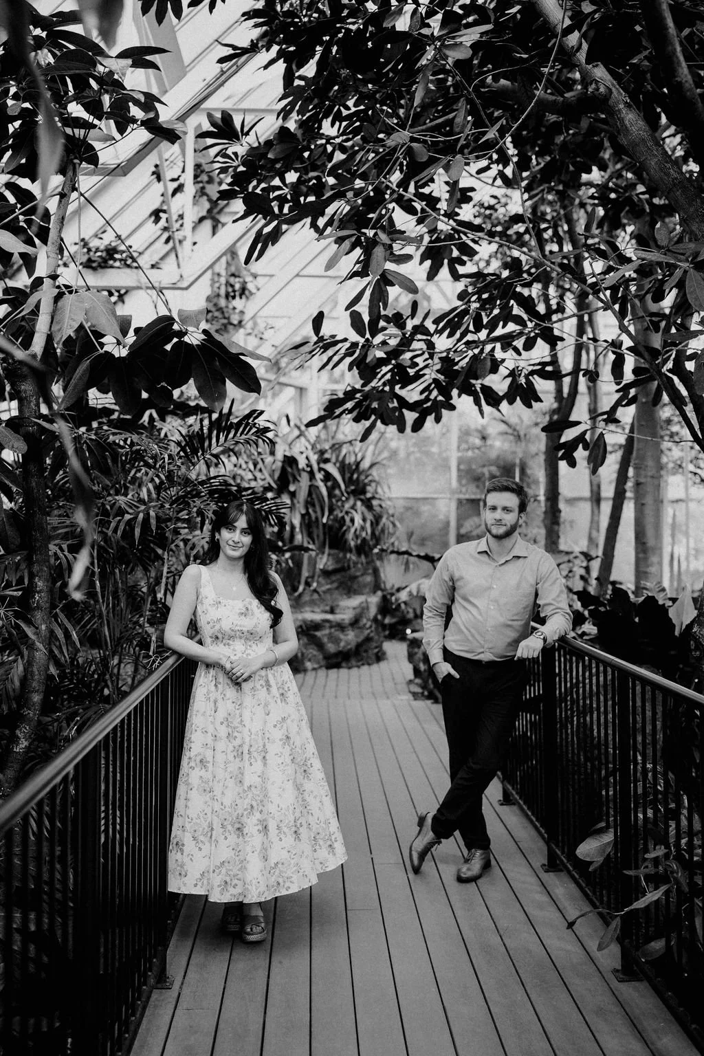 Engagement photo ideas for a relaxed pose, with a couple leaning on the black railings of a wooden greenhouse walkway in a black and white photo.