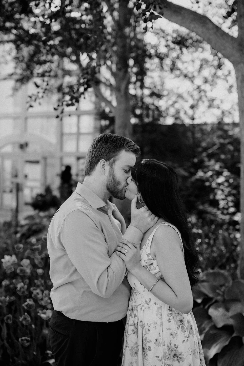Black and white photo of a couple sharing a kiss in a garden, with his hand on her cheek and her engagement ring in view.