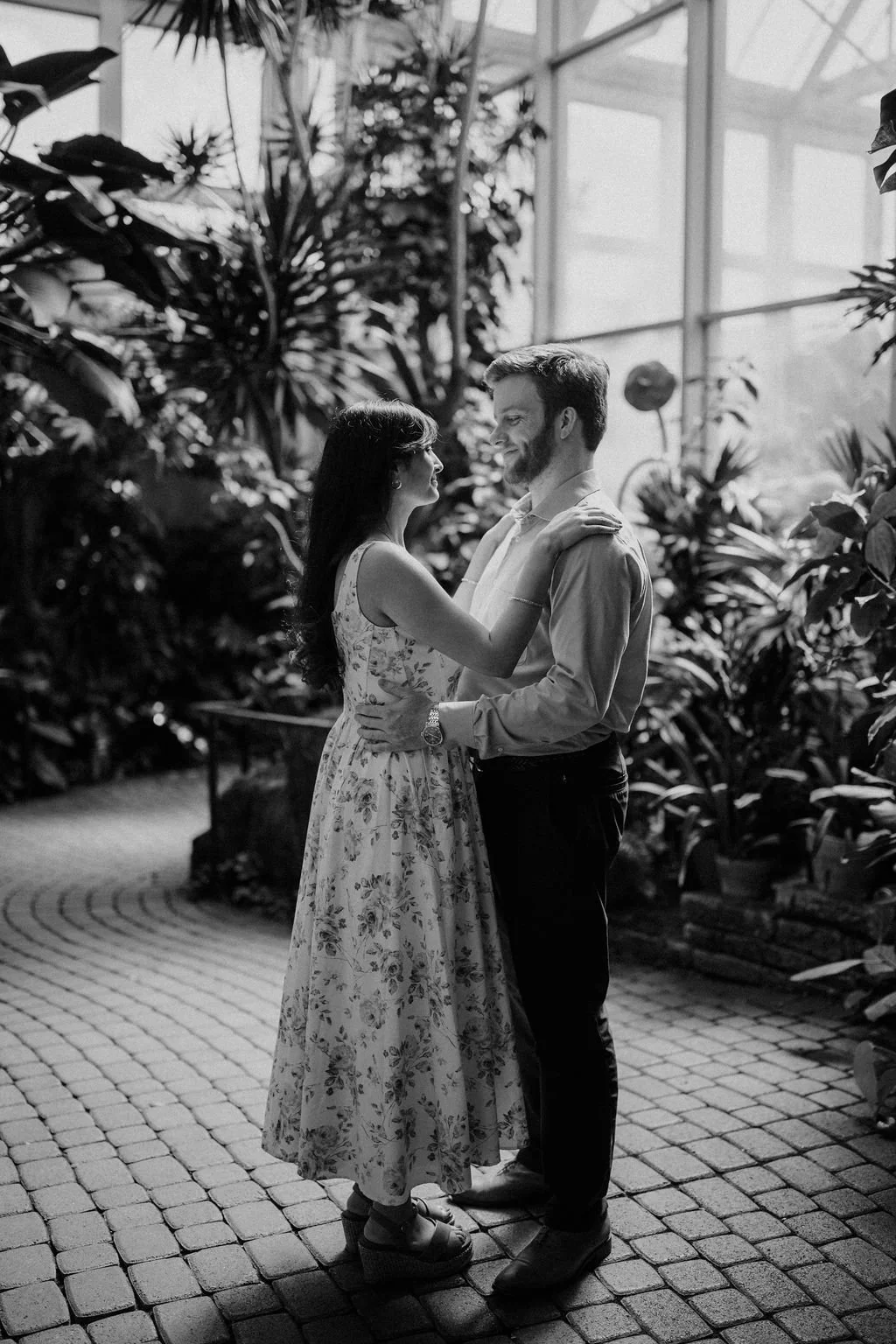 Engagement photo ideas in a botanical garden conservatory, with a couple facing each other in a black and white portrait while she rests her hand on his shoulder in a floral midi dress.