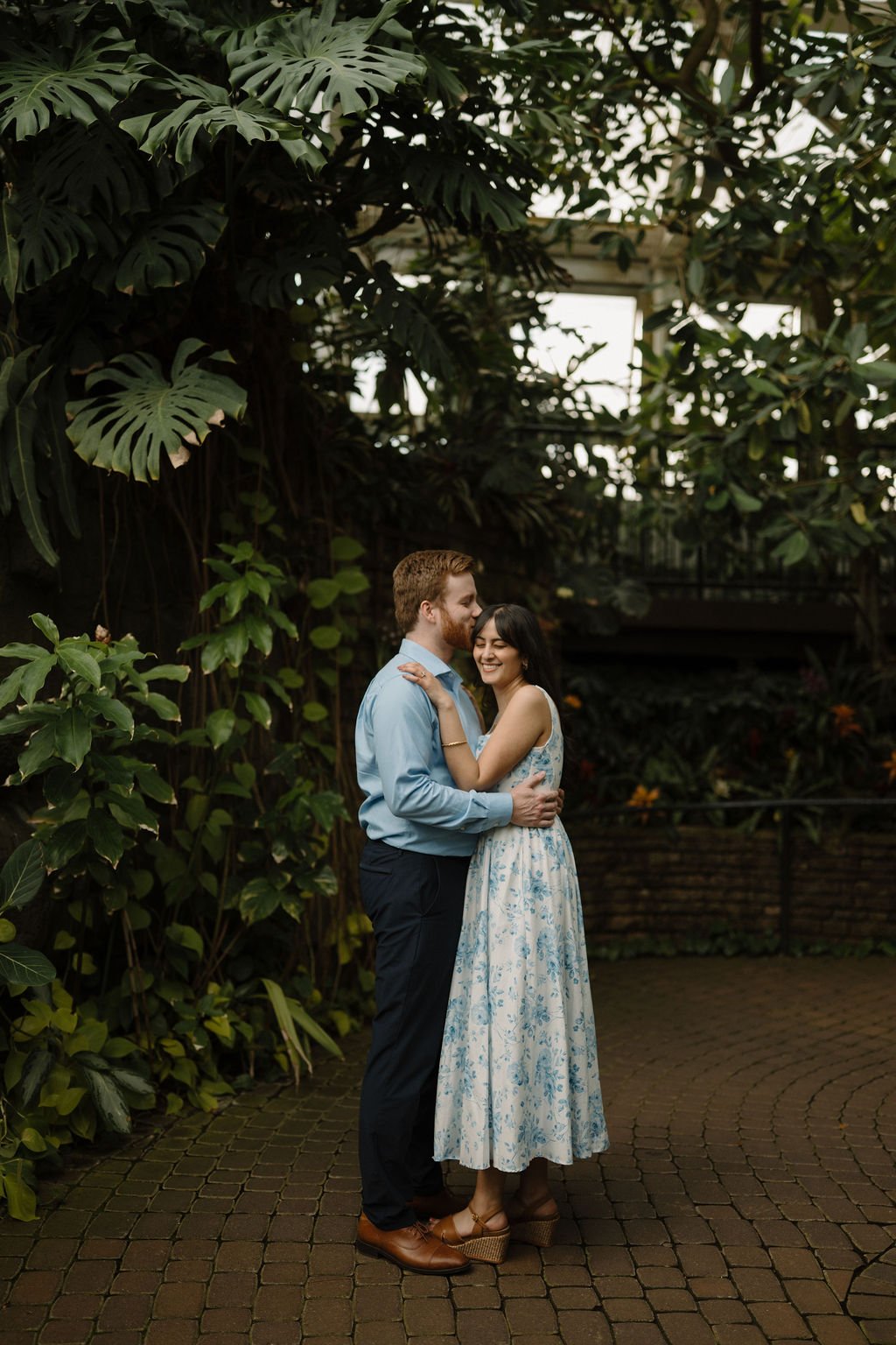 Smiling woman in a blue floral dress with her eyes closed as her fiancé kisses her forehead, surrounded by large monstera leaves inside a greenhouse.