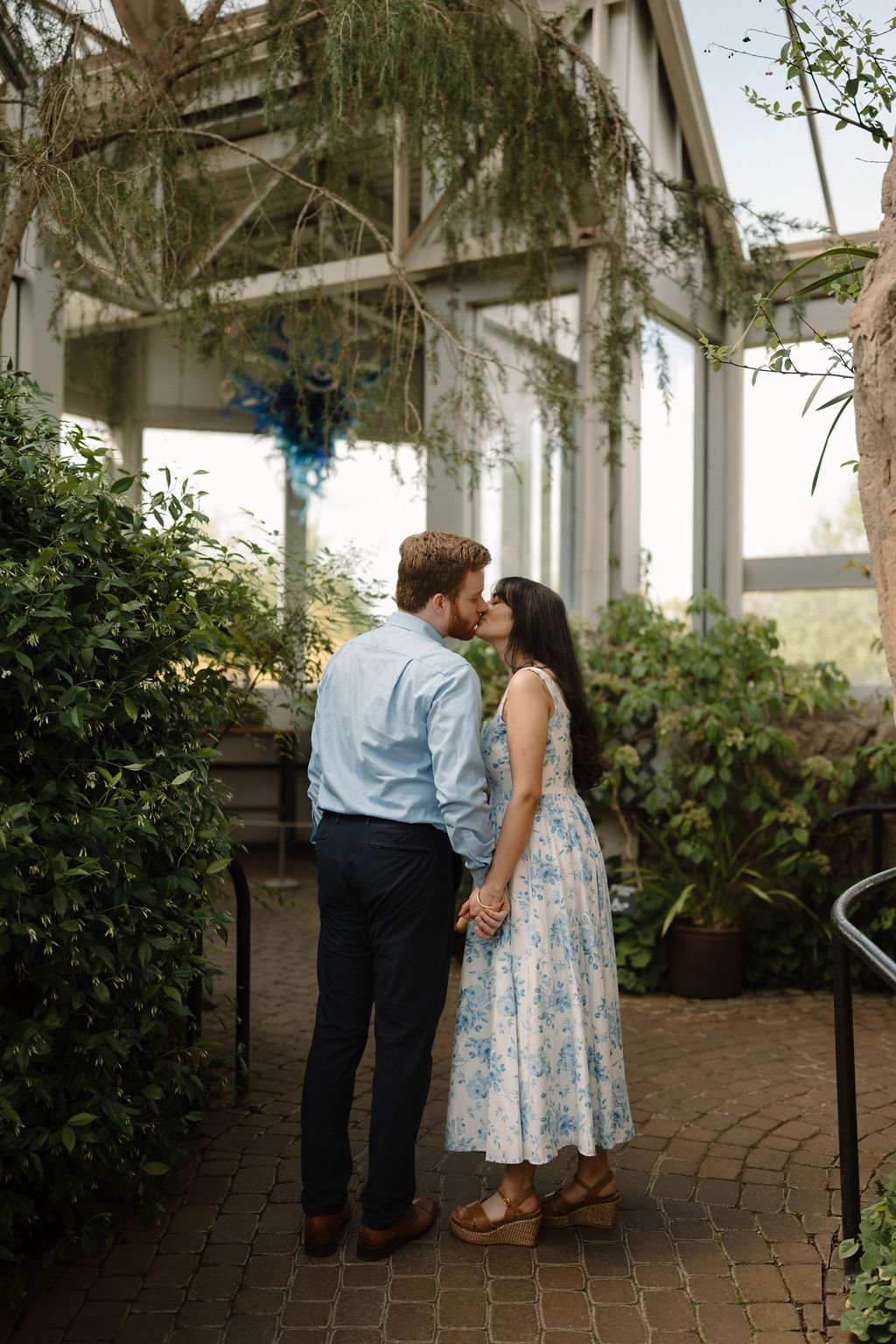 Couple sharing a kiss and holding hands on a brick path inside a greenhouse, with a hanging blue glass sculpture behind them.