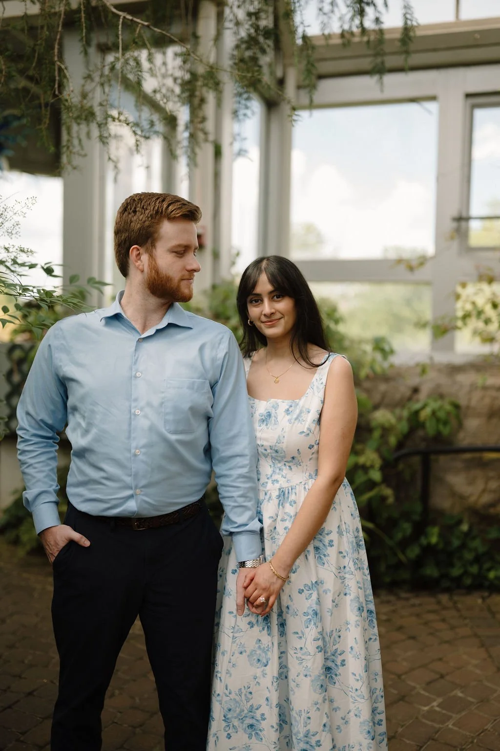 Woman in a blue floral dress smiling at the camera as her fiancé looks down at her, holding hands so her engagement ring shows.