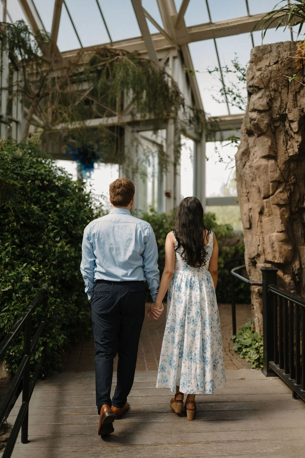 Engagement photo ideas with a couple walking hand in hand away from the camera along a wooden greenhouse walkway next to a large rock wall.